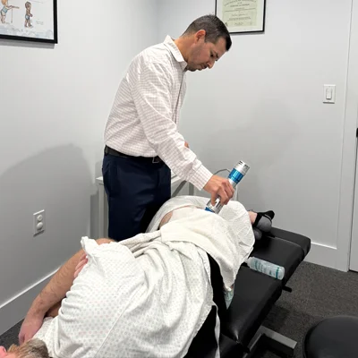 The chiropractor stands holding shockwave instrument against patient's leg wearing white