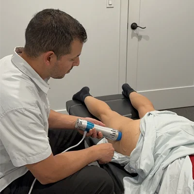 Overt the shoulder shot of chiropractor using the shockwave instrument on a patient's back upper thigh