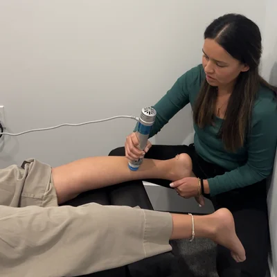 A patient has one pant leg rolled up while the chiropractor uses the shockwave instrument on the calf muscle